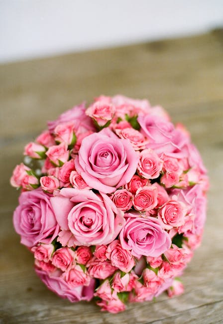 A round bouquet of fresh pink roses and smaller pink buds, arranged tightly in a compact, spherical shape. The roses have soft, layered petals with gentle curves, displaying a delicate pink hue, and a