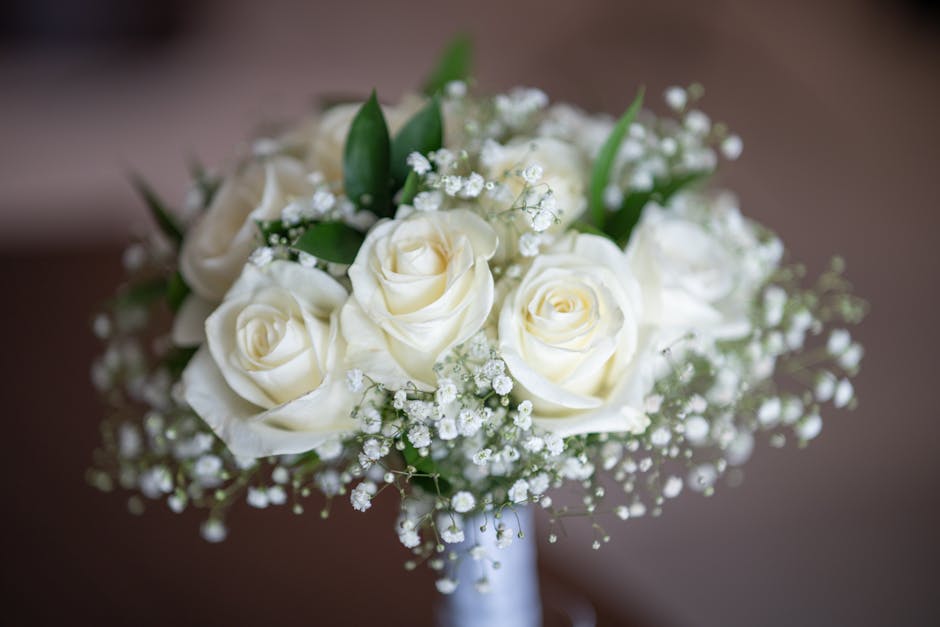 A floral arrangement featuring a bouquet of ivory-white roses with fully opened petals, complemented by sprigs of baby's breath and a few green leaves. The roses are arranged in a compact, rounded sty