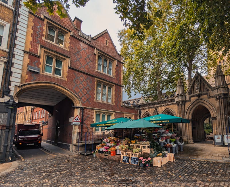 A flower stall set up on a cobbled street with an arched stone building in the background, featuring a variety of fresh flowers in vibrant colors including pinks, reds, yellows, and whites. The flower