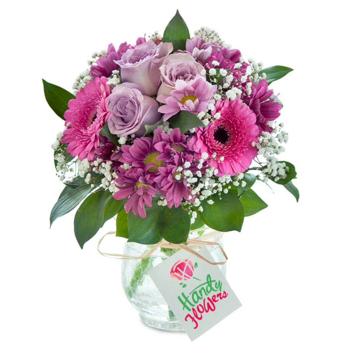 Set before a crisp white backdrop, this spectacular bouquet from a Twickenham florist captures the region's vibrant creativity. Soft lavender roses, their petals gracefully unfolding, form the gentle heart of the arrangement-suggestive of the sophisticated, soothing character that lingers in the borough's historic gardens. They are embraced by vivid hot-pink Gerbera daisies, their broad petals tinting the air with cheerful colour and radiating positivity akin to a sunlit stroll near York House Gardens. Dark magenta chrysanthemums cluster around, each dotted with a subtle, grassy centre, weaving a striking layer of texture through the bouquet. Generous sprigs of white baby's breath, finely branched and feather-light, introduce a play of airiness and clarity, allowing the ensemble to breathe softly. Glossy leaves in deep green surround the entire display, offering harmony and completion. A rounded glass vase with an intricate crackled surface amplifies the atmospheric light that falls across the blooms, lending rustic elegance reminiscent of a well-loved Twickenham café. Tied with rustic raffia, a stark white tag sways gently, ready for a hand-written note. Perfectly suited for anniversaries, birthdays, or expressions of gratitude, this bouquet speaks to both celebration and sincere gesture, reliably available for same-day local delivery in Twickenham and surrounding riverside districts.